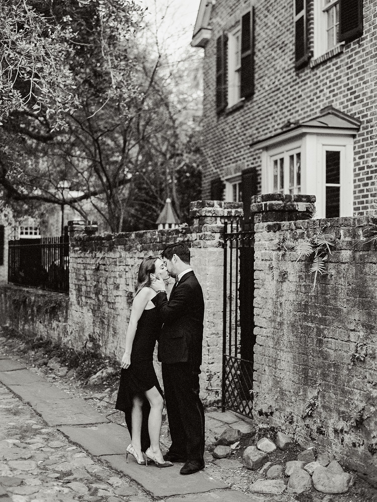 Man in tuxedo kissing woman in black dress while standing in cobblestone alleyway in Charleston
