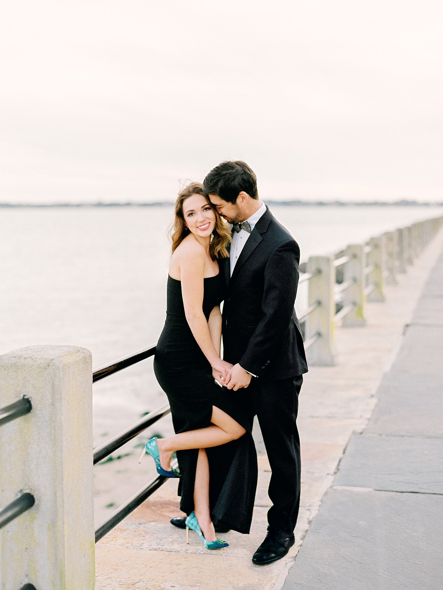 Man in tuxedo and girl in black dress holding hands and laughing on by water during Charleston Couple session