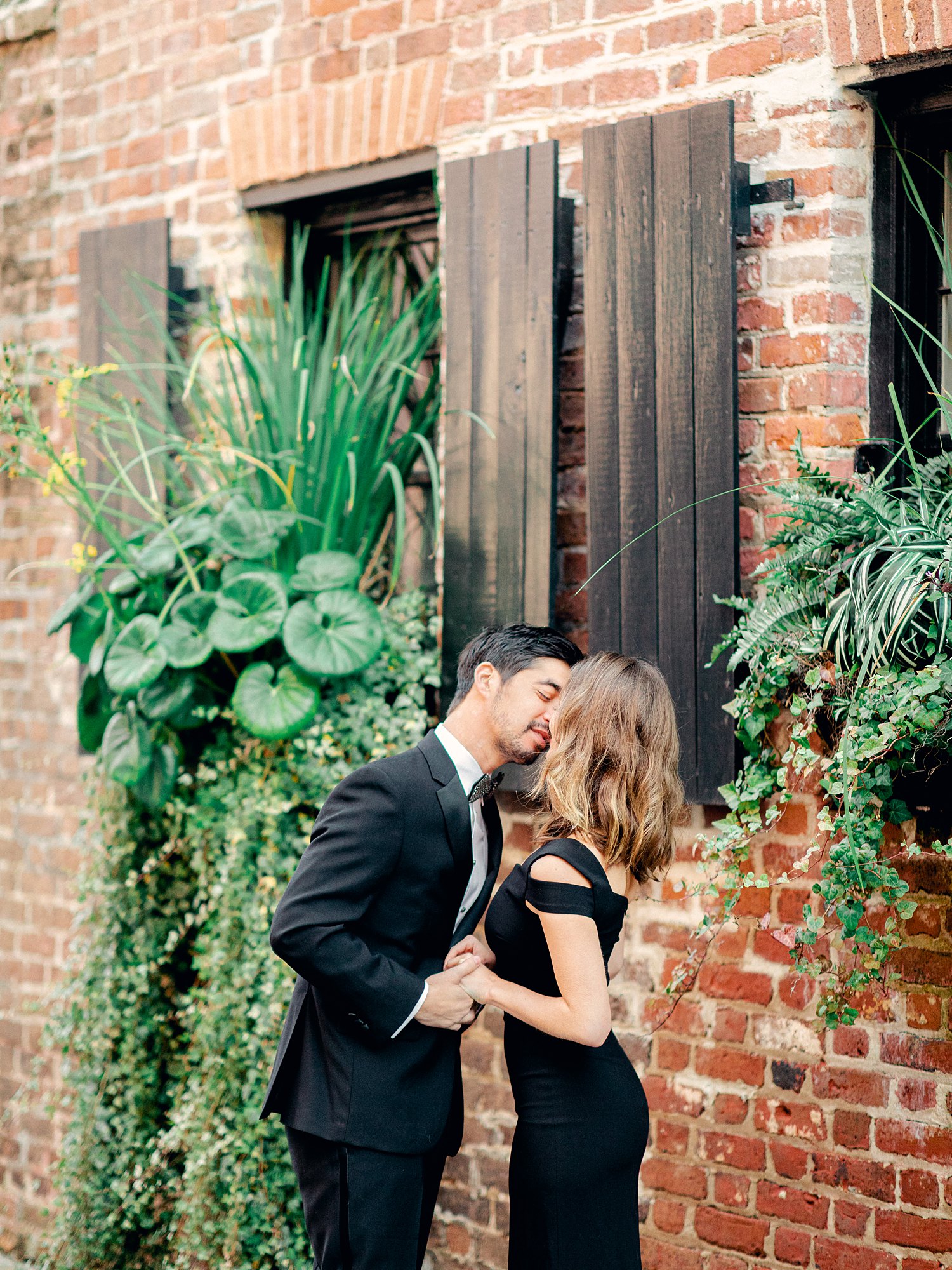 Man in black tuxedo kissing girl in black dress in front of red brick and green plants