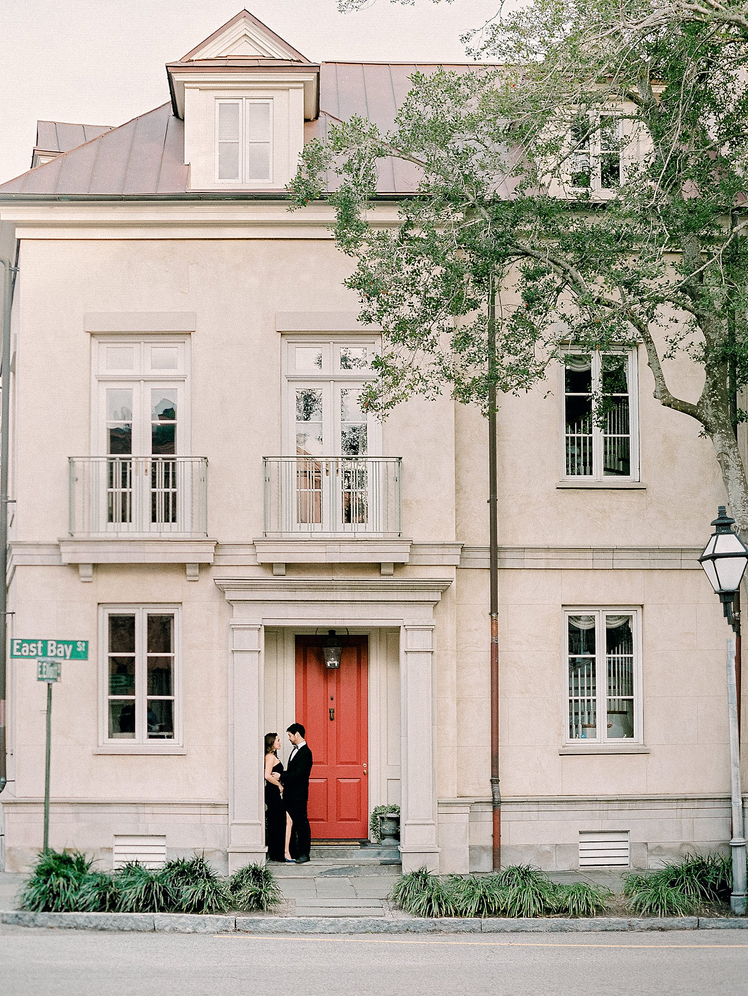 Man in tuxedo hugging girl in black dress by orange door on doorstep of townhome in Charleston engagement