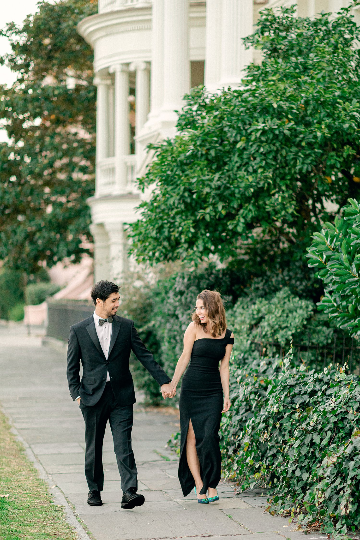 Couple laughing and holding hands dressed in black on green treelined sidewalk during Charleston Engagement