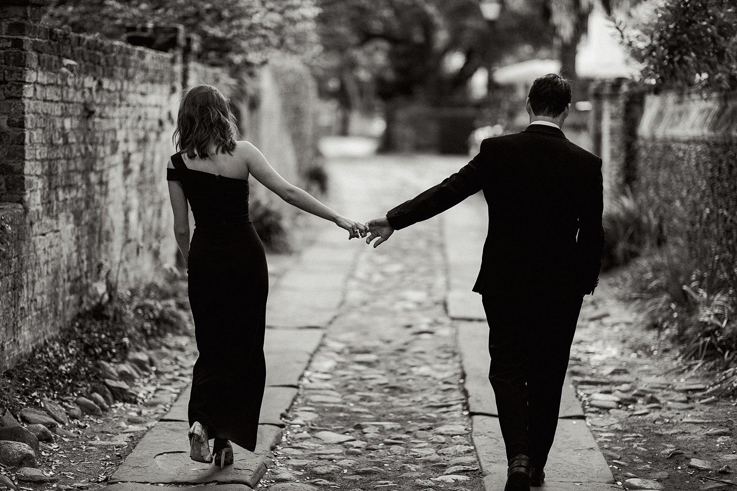 Man in tuxedo holding hands with woman in black dress walking down cobblestone alley and brick wall