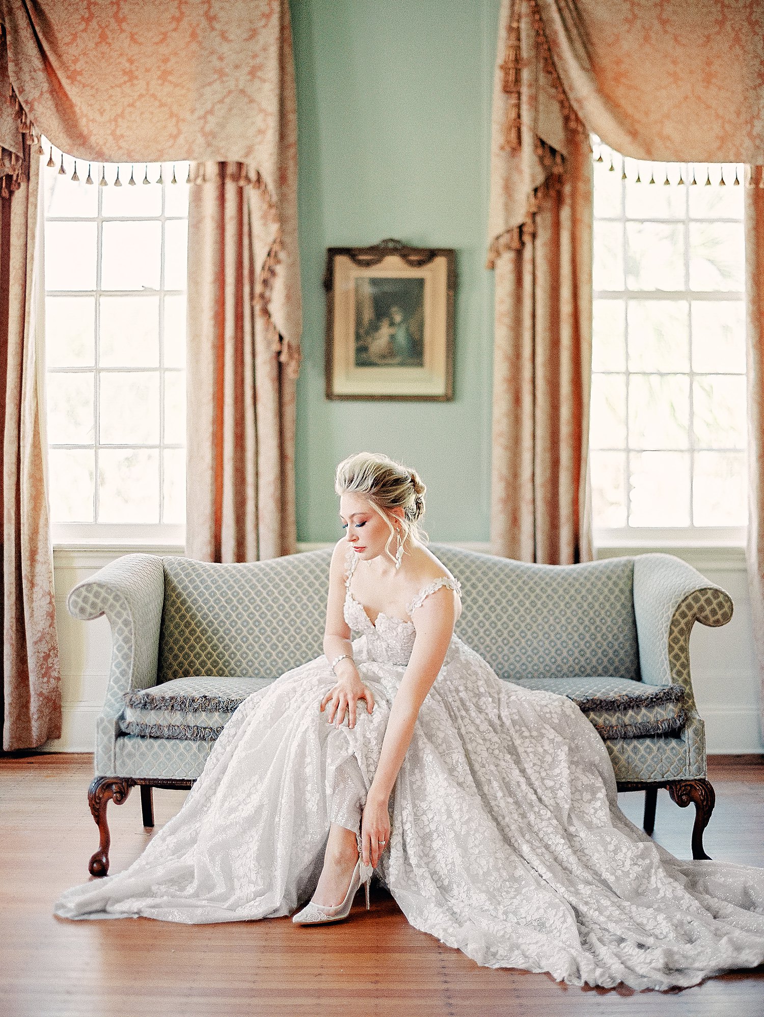 bride in white wedding dress putting on shoes seated on green couch at Lowdnes Grove Charleston