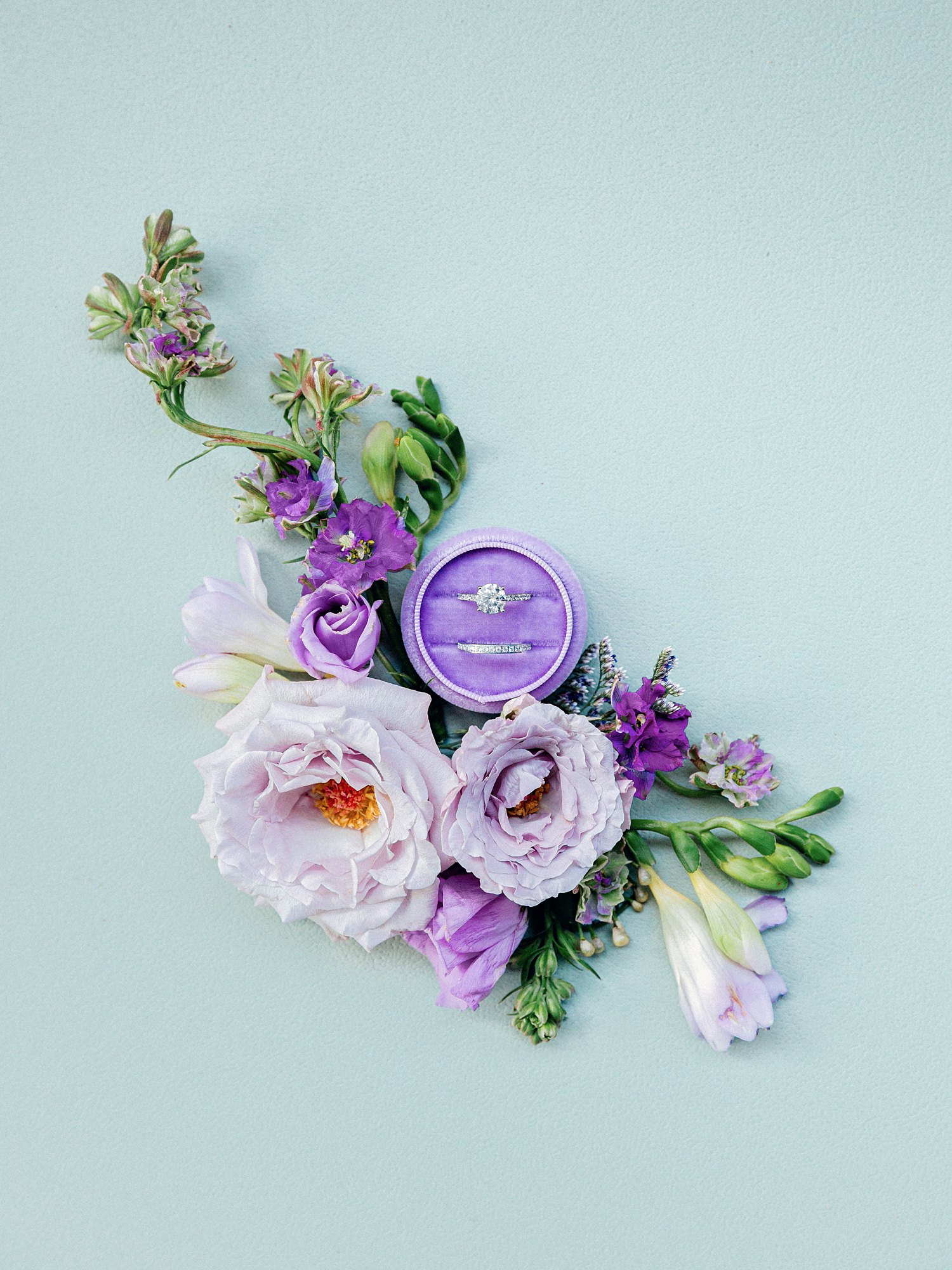 diamond engagement ring and wedding band in round purple ring box sitting among purple flowers against a green background flat lay 