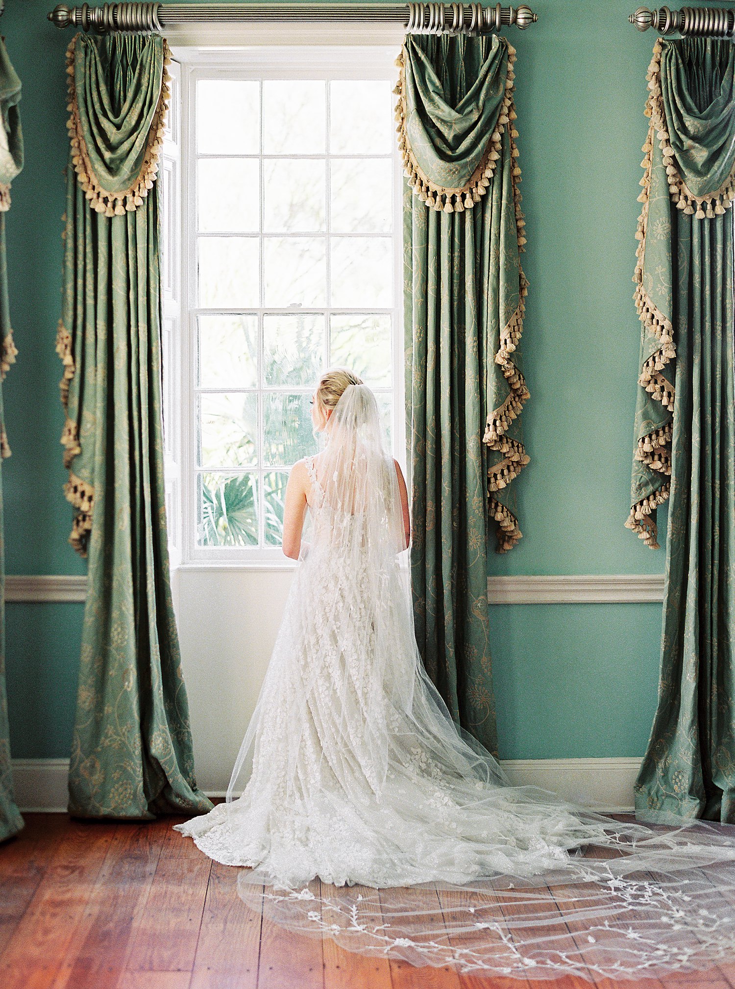 Bride in white wedding dress standing at window with green curtains at Lowndes Grove