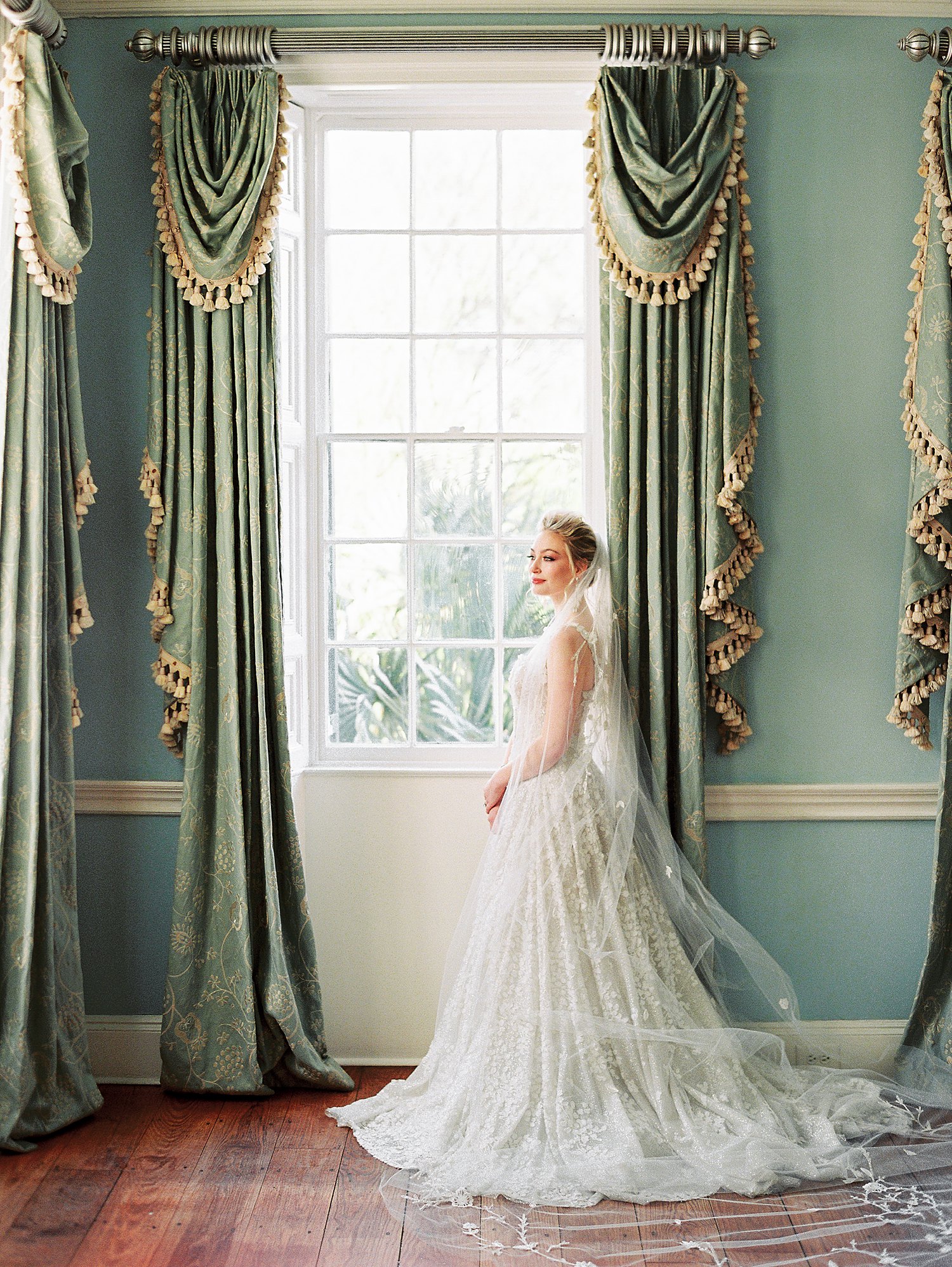 Bride in white wedding dress and veil standing at window with green curtains at Lowndes Grove Charleston