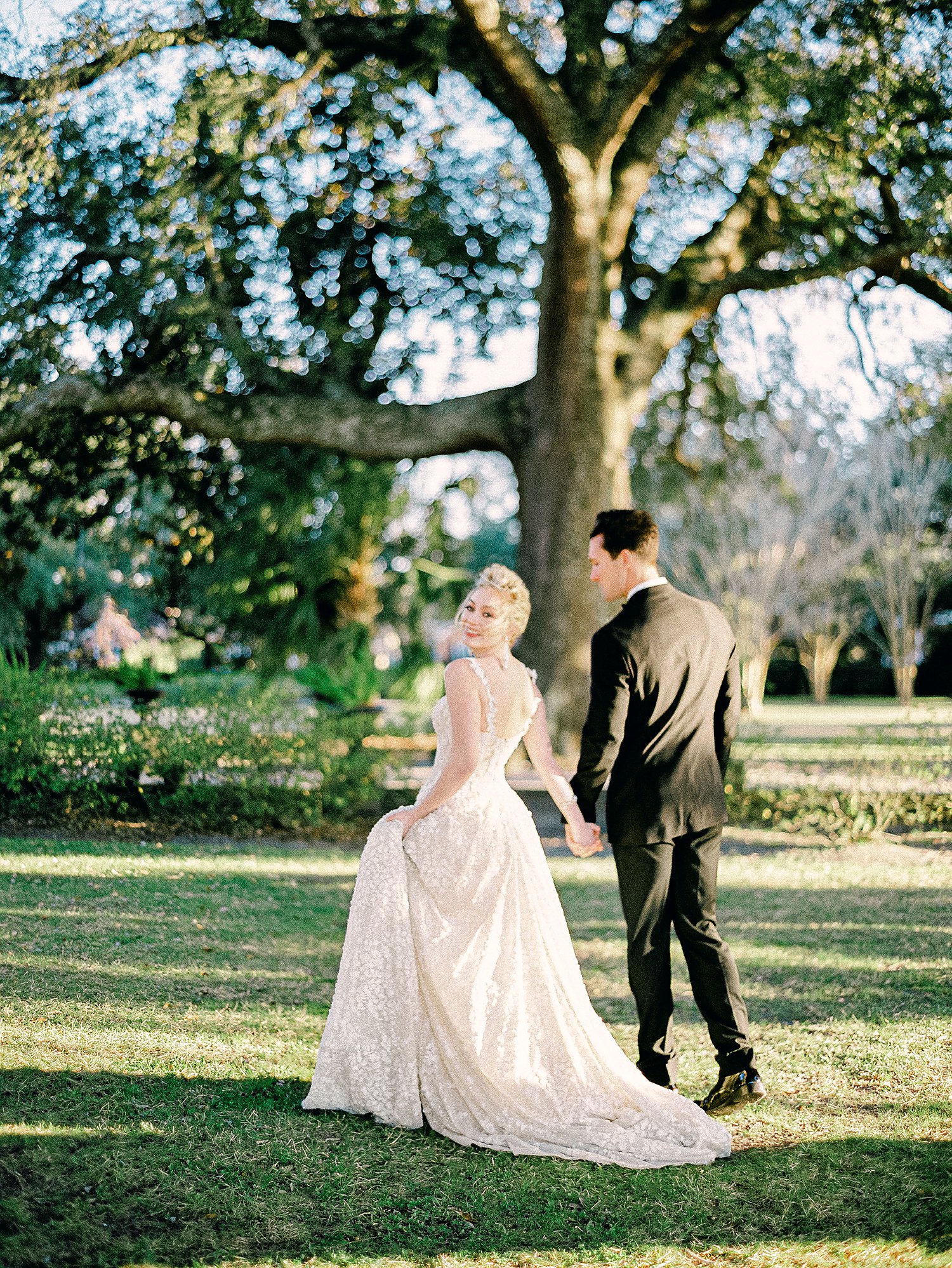 bride in white wedding dress walking with groom in black tuxedo in green garden