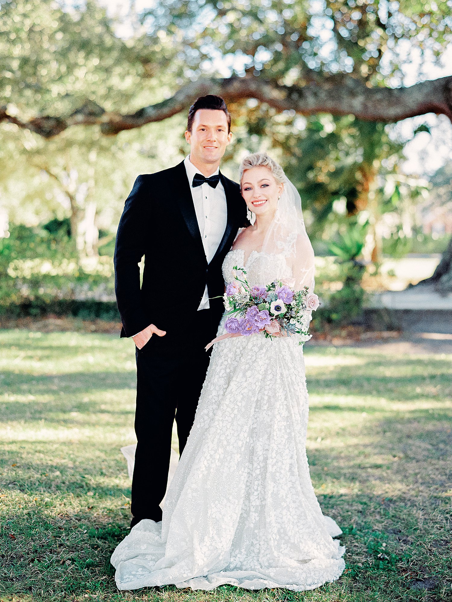 bride in white dress and purple floral bouquet standing with with groom in black tuxedo in green garden at Lowndes Grove Charleston wedding