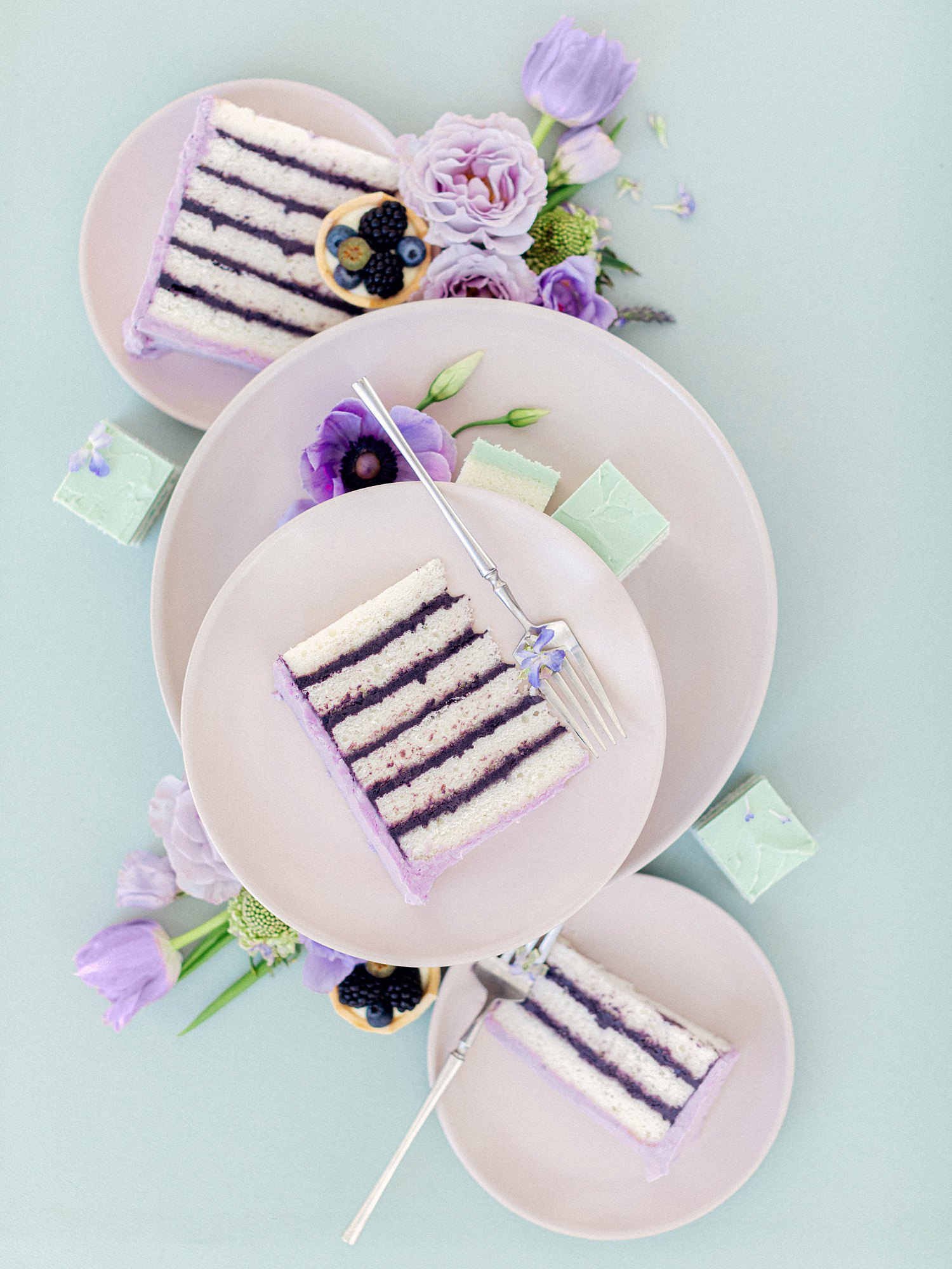 purple cake slices laying on plates surrounded by purple florals against green background wedding flat lay
