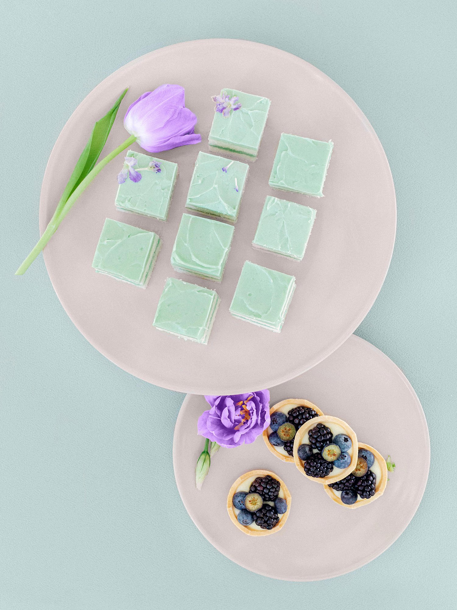 green cake squares and berry tarts on plates with purple florals against green background wedding flat lay