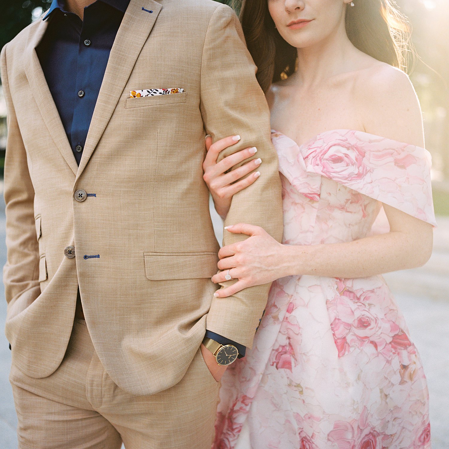 Woman in Pink floral gown hand and arm around man in tan suite with gold watch downtown dallas engagement