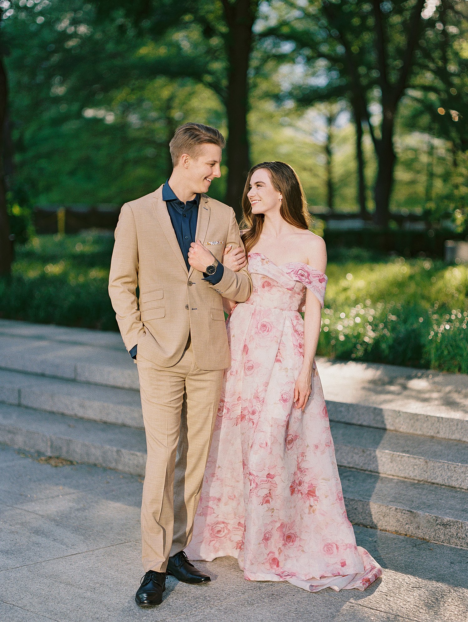 smiling woman in Pink floral gown standing with man in tan suite and blue shirt dallas garden engagement