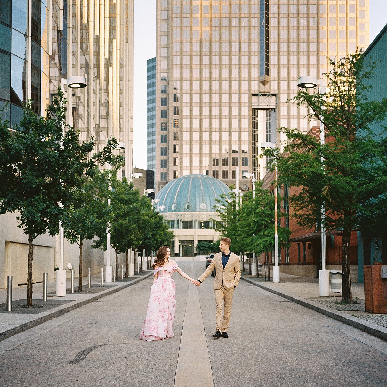 woman in Pink floral gown holding hands with man in tan suite downtown Dallas street