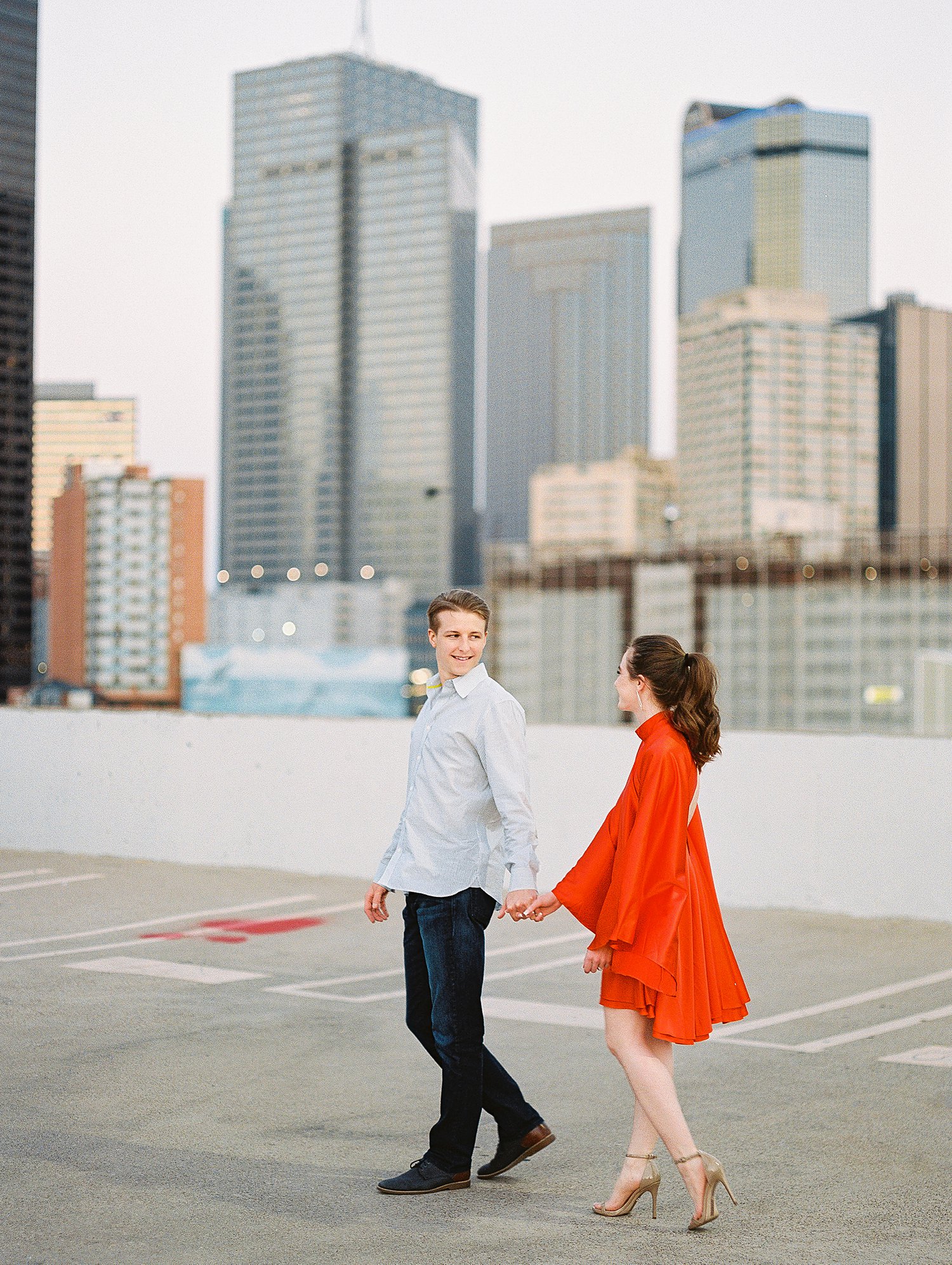 woman in orange dress and pony tail walking with smiling man downtown Dallas engagement session