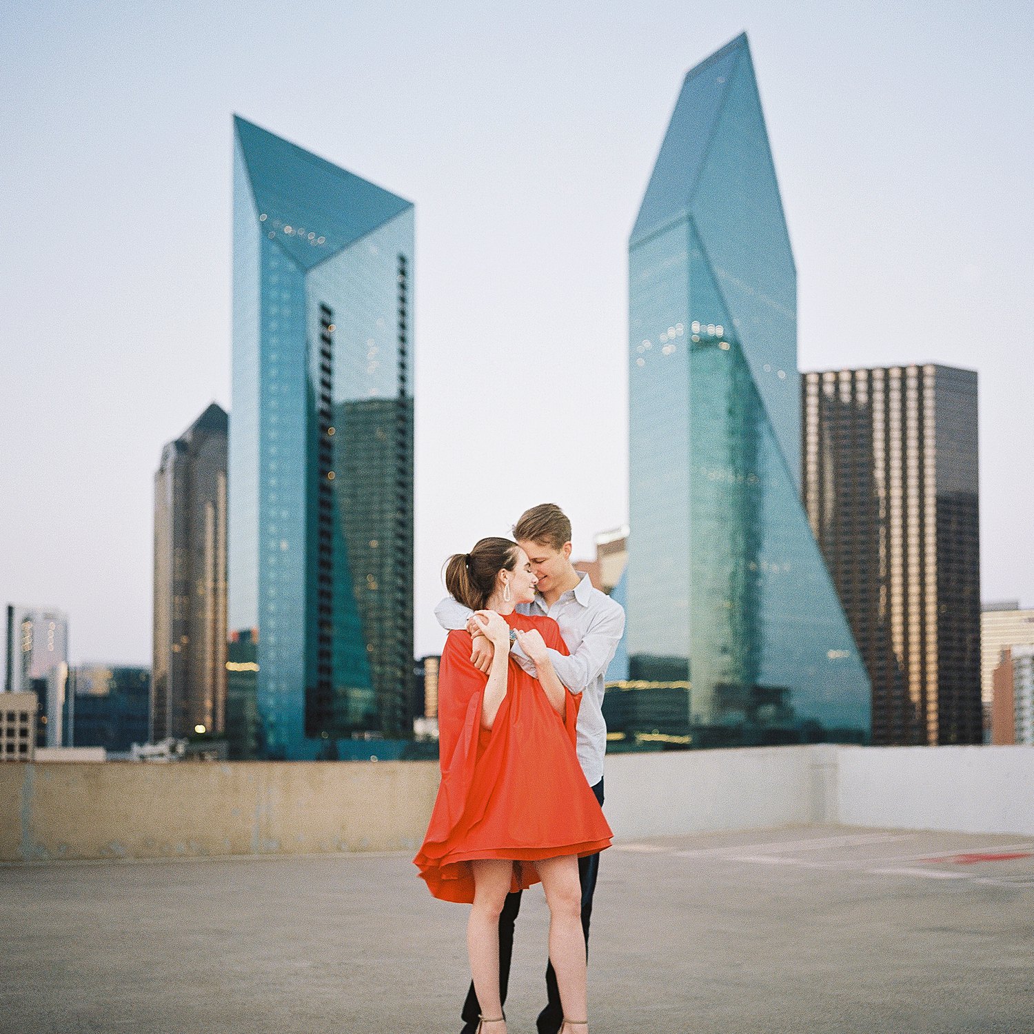 woman in orange dress and pony tail embraced by man on downtown Dallas rooftop engagement session