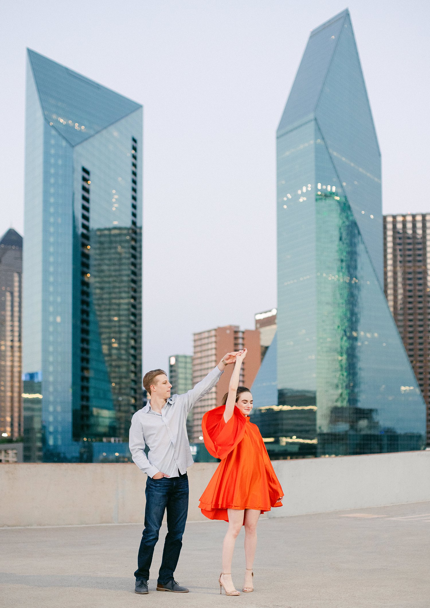 woman in orange dress and pony tail twirling with man on downtown Dallas rooftop engagement session