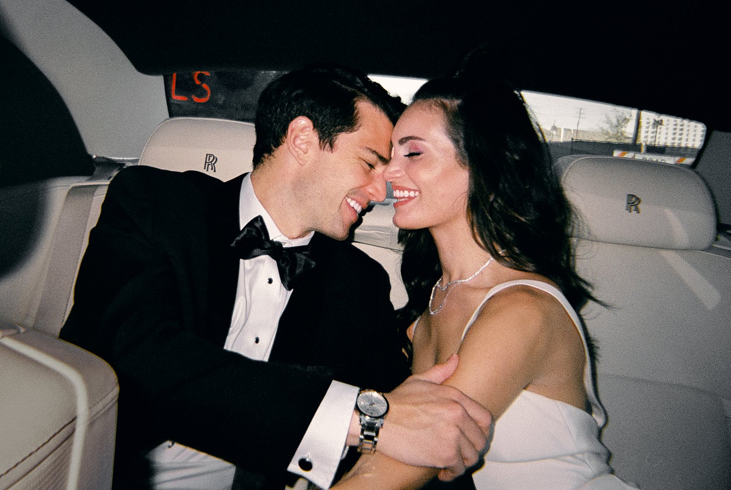 man in black tuxedo with woman in white dress sitting in car smiling