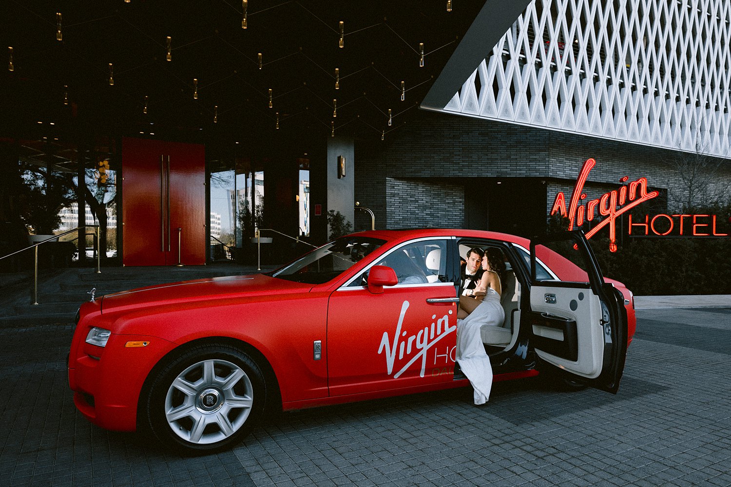 man in black tuxedo with woman in white dress sitting in red Virgin hotel Dallas Rolls Royce car 