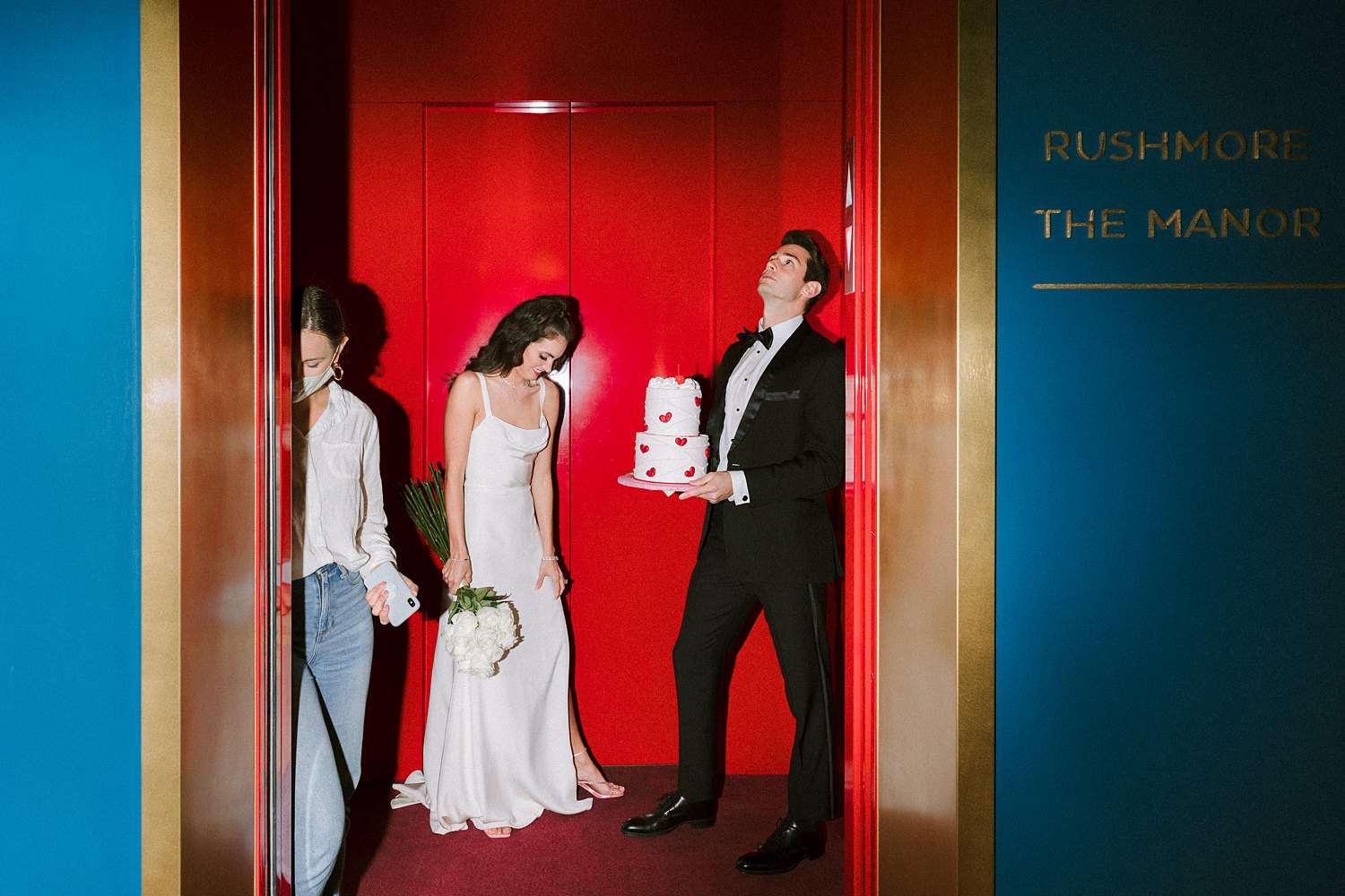 Man in black tuxedo and bowtie standing with cake in hands next to woman in white dress holding flower bouquet in red elevator at Virgin Hotel Dallas