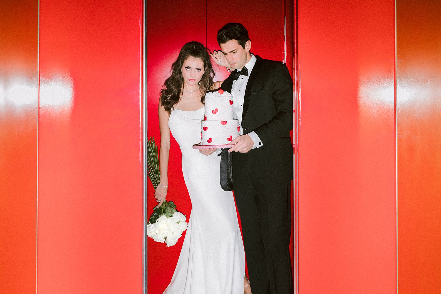 Man in black tuxedo standing with cake in hands next to woman in white dress holding flower bouquet in red elevator