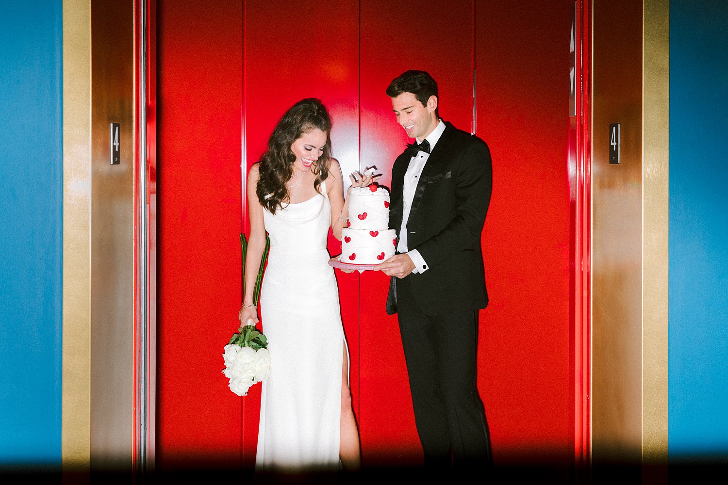 Man in black tuxedo and bowtie standing with Vintage wedding cake in hands next to woman in white dress holding flower bouquet in red elevator with blue walls