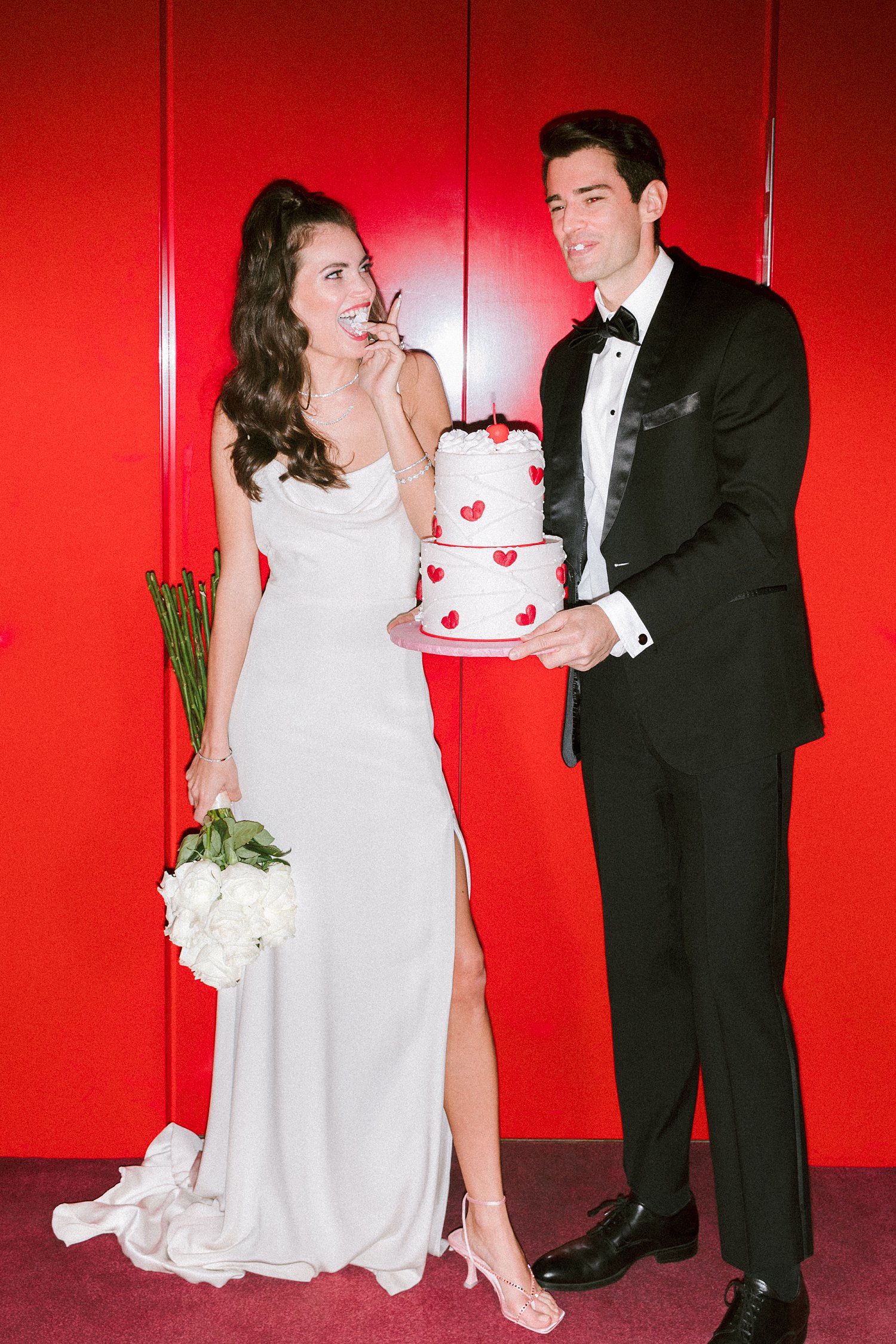 Man in black tuxedo and bowtie standing with cake in hands next to laughing woman in white dress holding flower bouquet in red elevator Virgin Hotel Dallas
