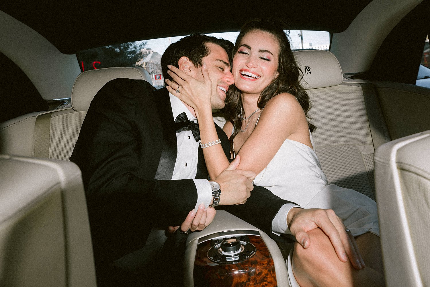 man in black tuxedo with woman in white dress sitting in car laughing