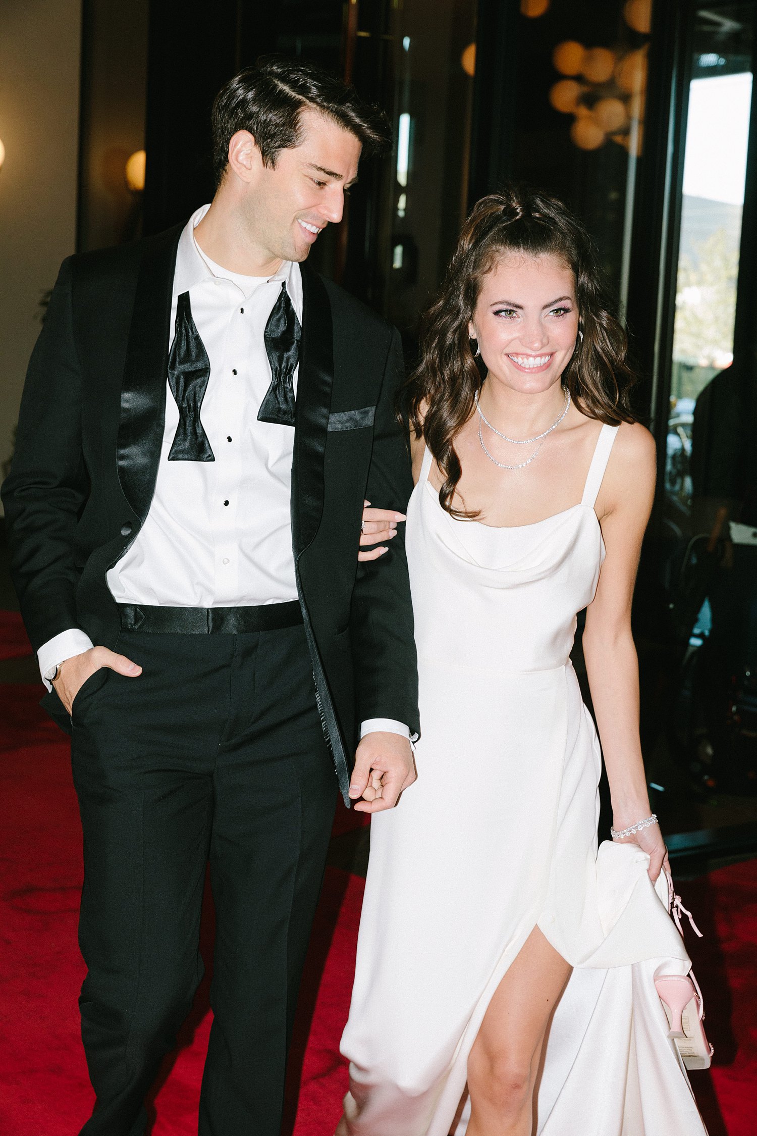 man in black tuxedo with woman in white dress walking in red carpet Virgin Hotel Dallas lobby smiling