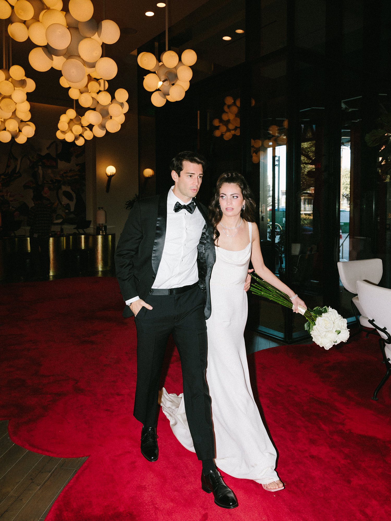 man in black tuxedo with woman in white dress walking in red carpet hotel lobby of Virgin Hotel Dallas