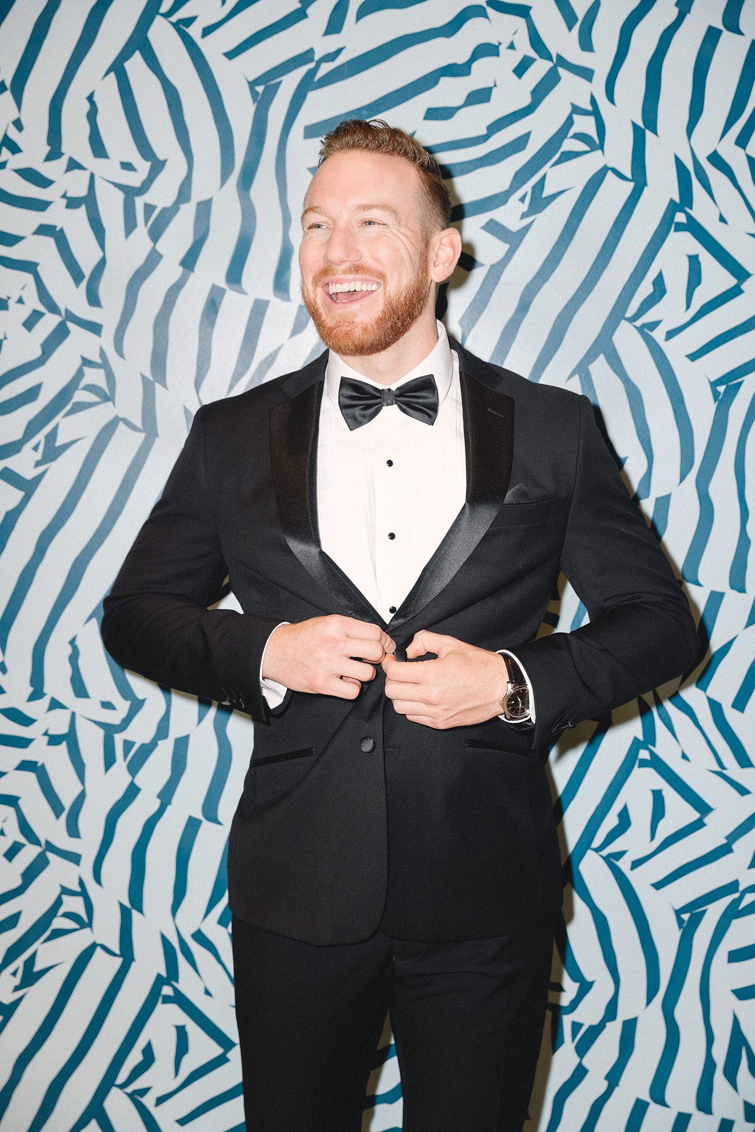 man in black tuxedo laughing in front of blue and white striped wall