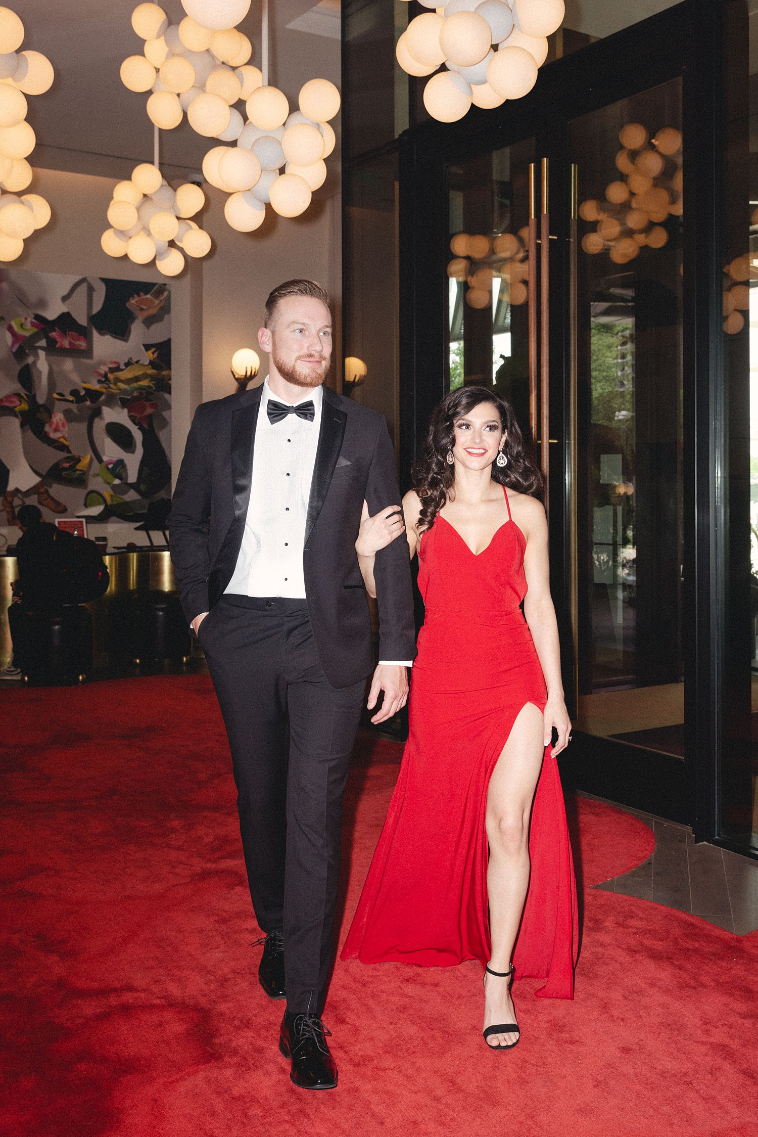 Woman in Red Dress and man in black tuxedo walking together into Virgin Hotel Dallas Engagement