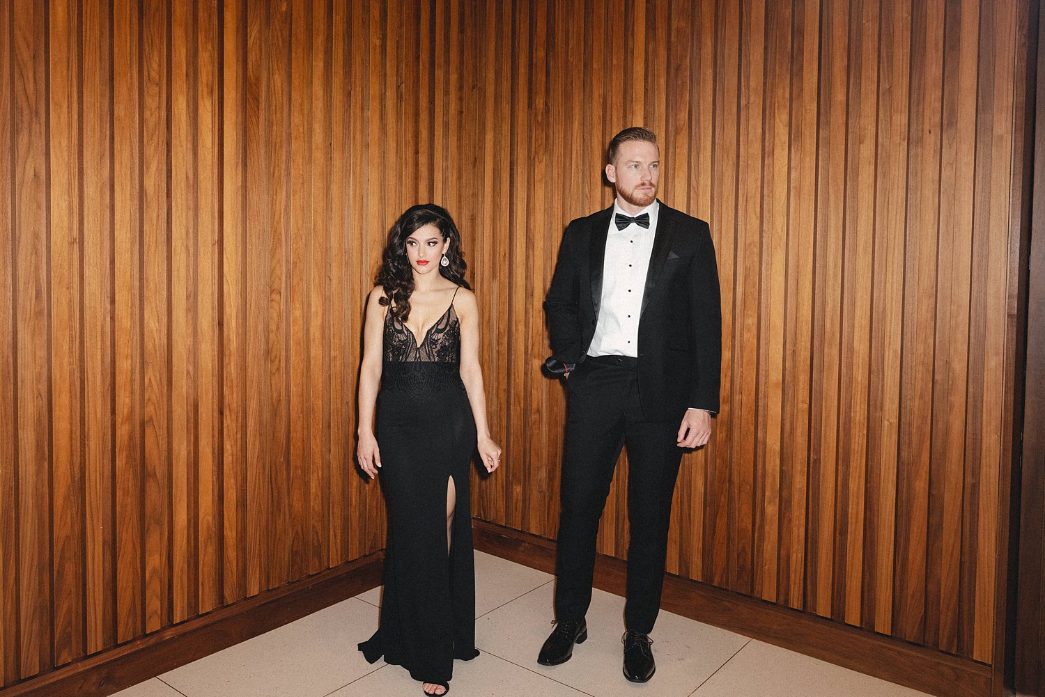 Woman in black dress and man in tuxedo with bow tie standing in front of wood paneled wall