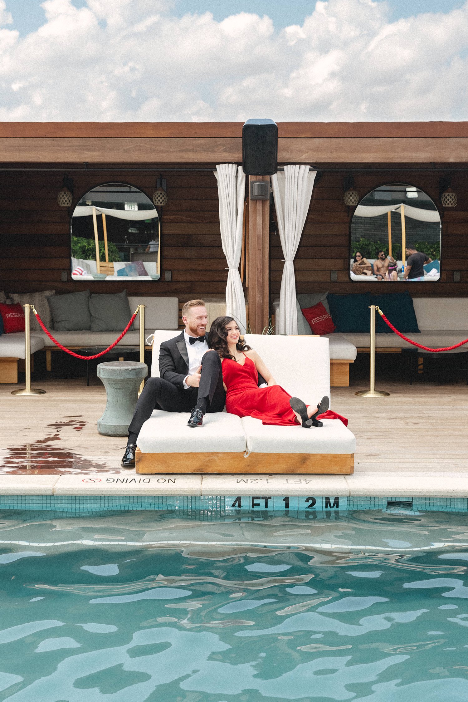 Woman in Dress sitting with man in tuxedo on white poolside deck sofa at Virgin Dallas Hotel engagement