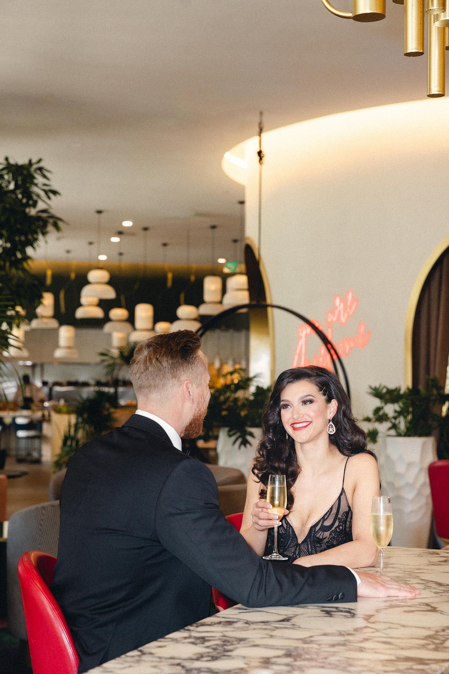 Woman in black dress and man in tuxedo toasting at bar at Virgin Hotel Dallas