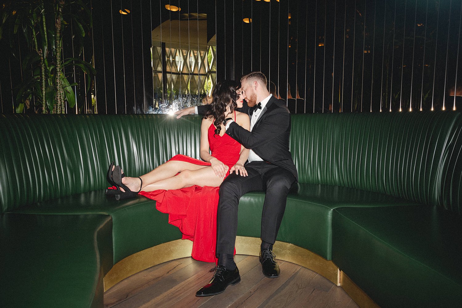 Woman in red Dress and man in black tuxedo sitting together in green booth at Virgin Hotel Dallas Engagement