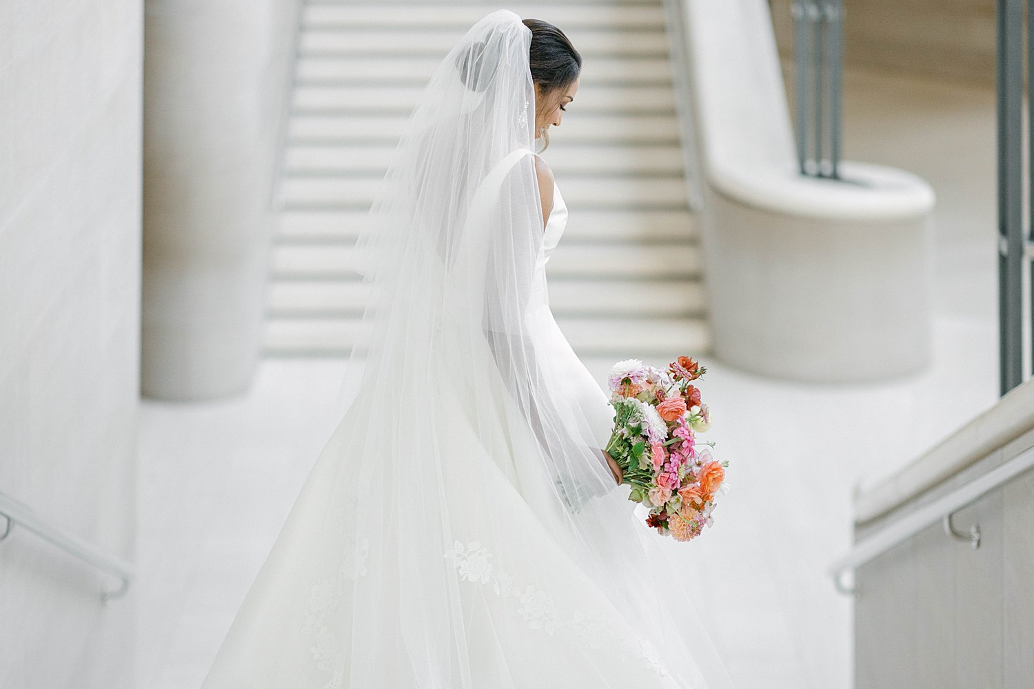 Bride in white wedding dress and veil holding colorful floral bouquet on top of staircase