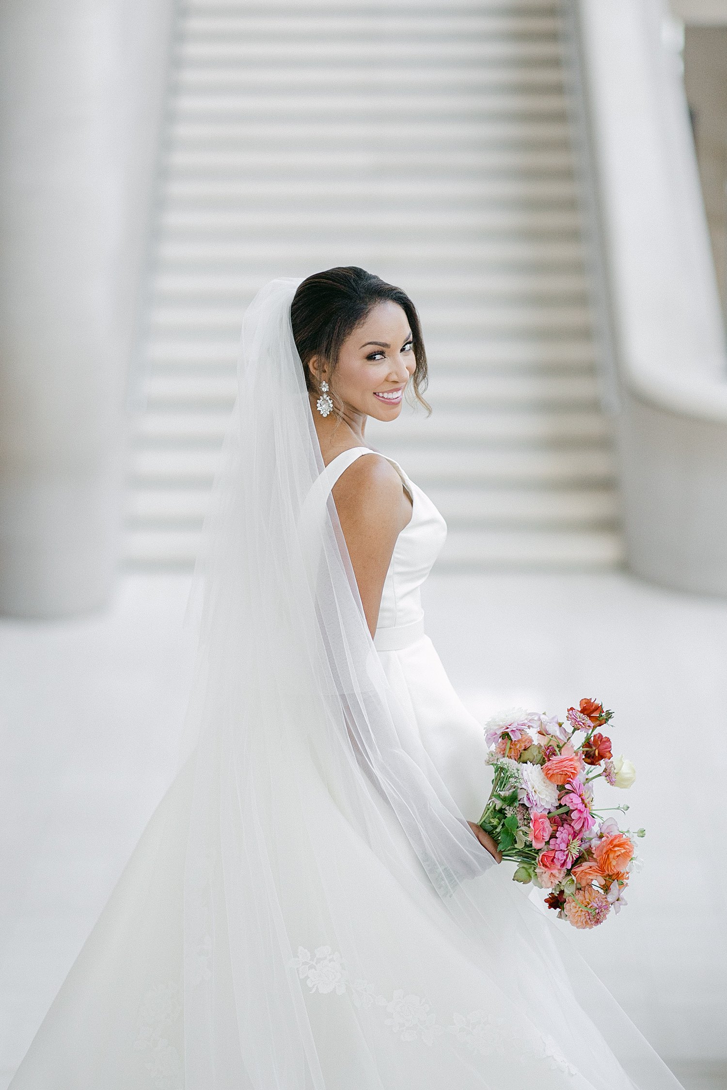 Bride in white wedding dress and veil holding colorful floral bouquet on top of staircase smiling Dallas Arts District
