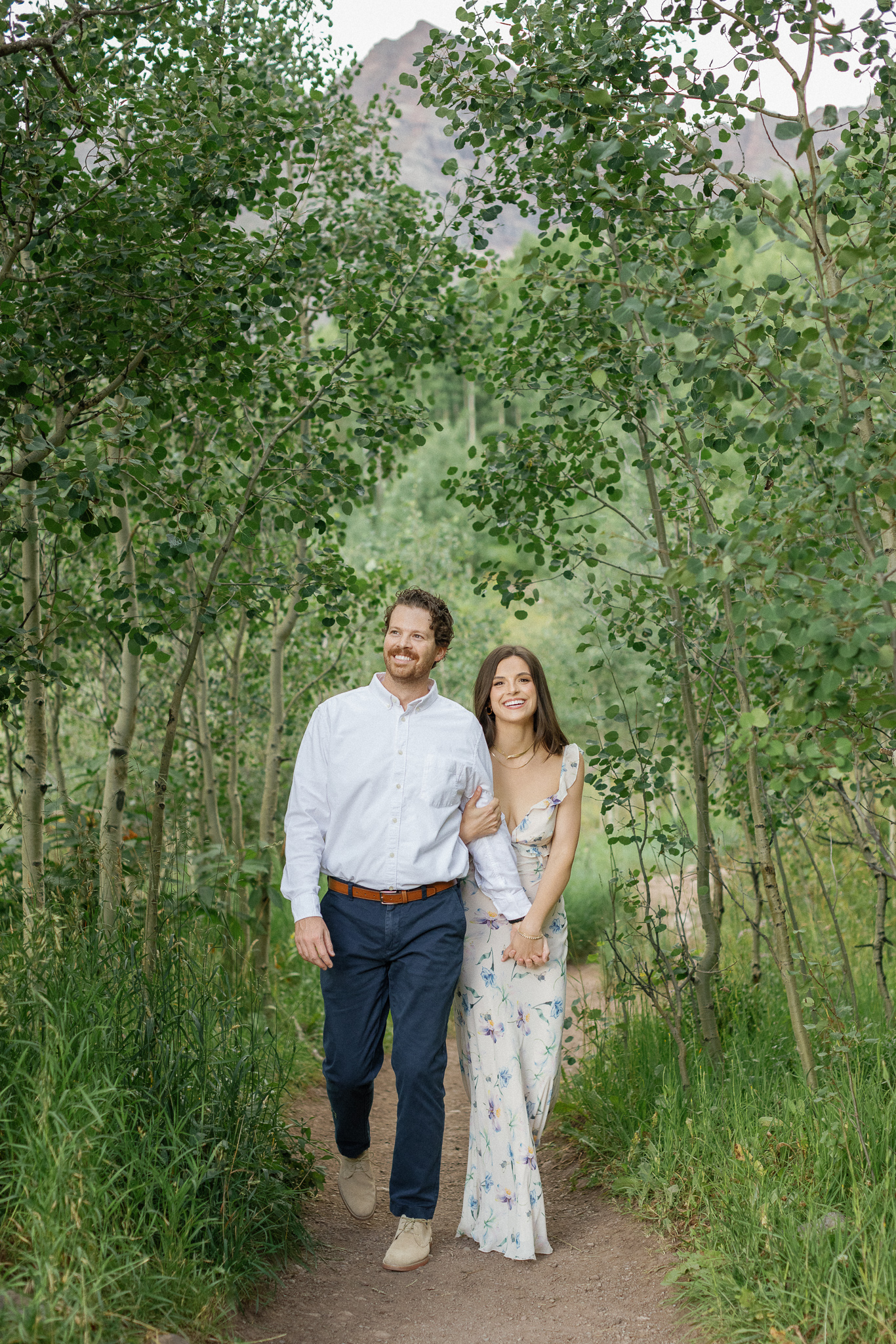 Man in white shirt and navy pants walking with smiling woman in white floral dress in a green aspen grove