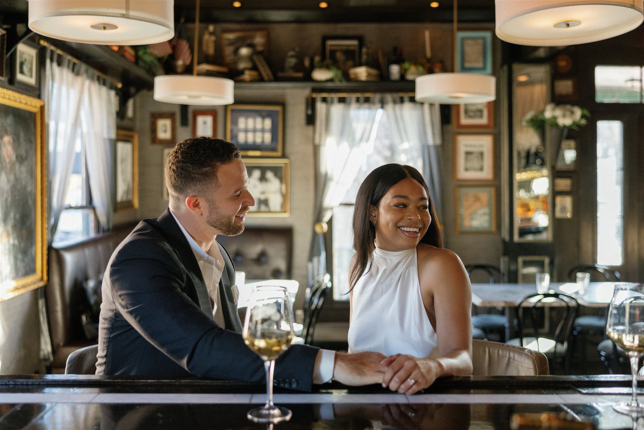Man in suit and woman in white dress sitting at bar holding hands smiling