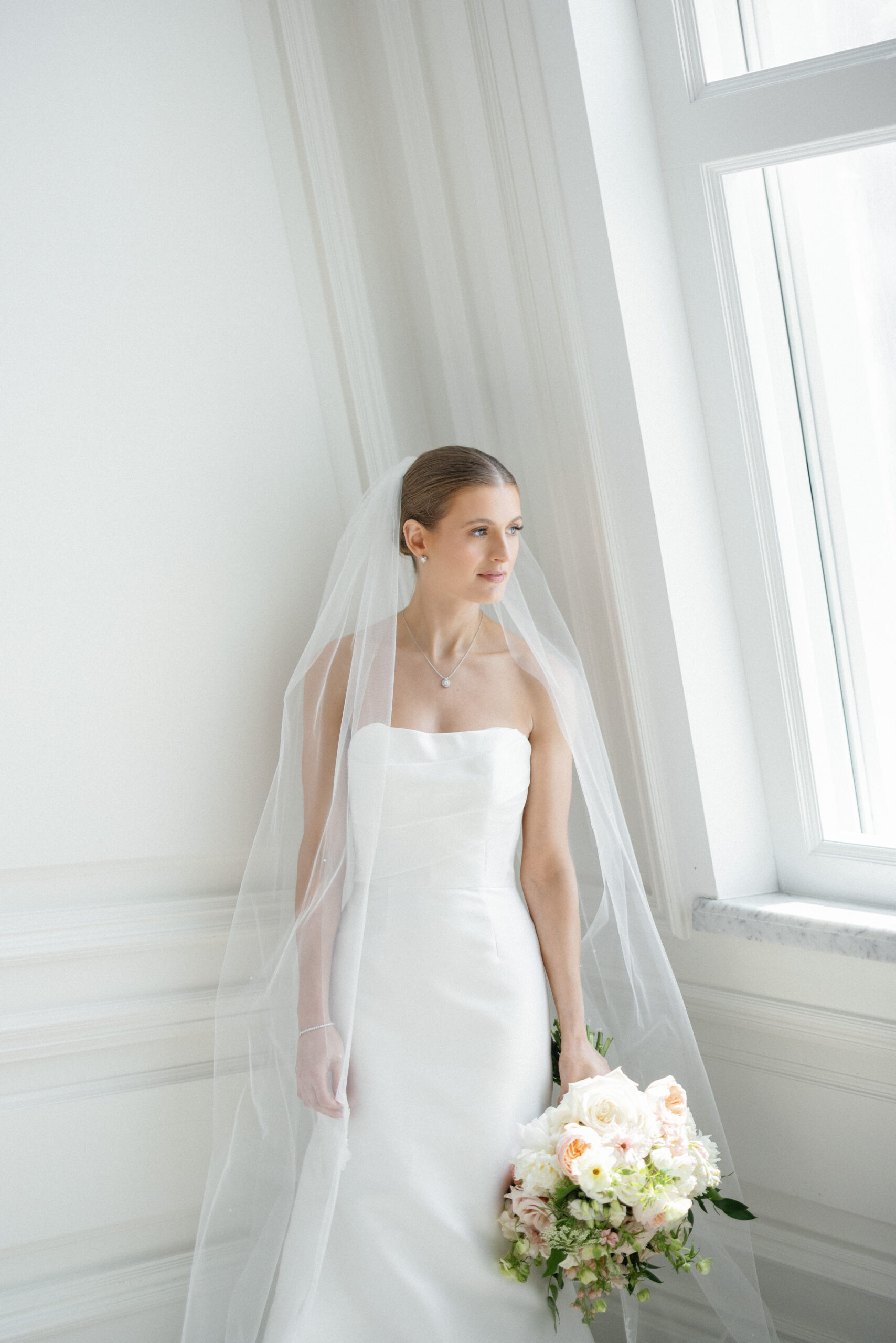 bride wearing strapless white wedding dress and long veil holding floral bouquet and looking out large bay windows at the Adolphus Hotel in Dallas